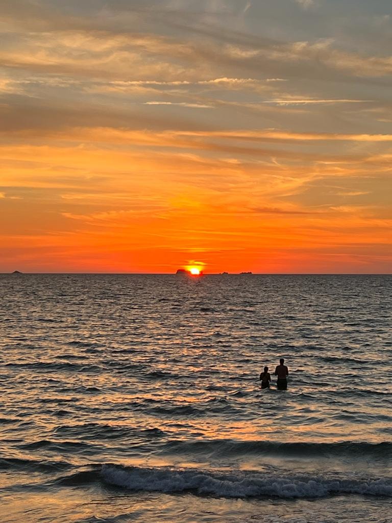 Two figures standing in the sea watching the sun set over the horizon
